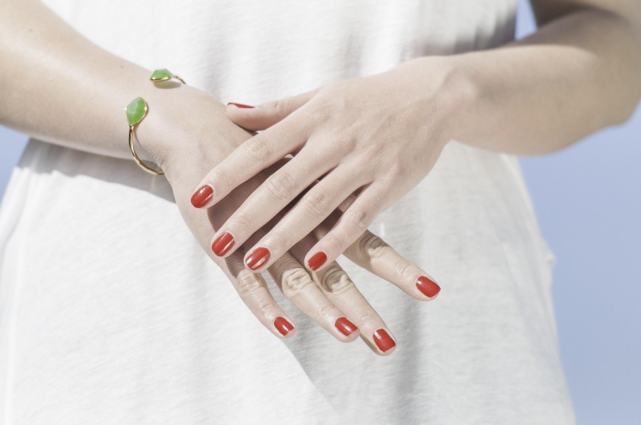 Close-up of a woman's crossed hands with neatly painted glossy red nails and a gold bracelet with green stones, against a white top.